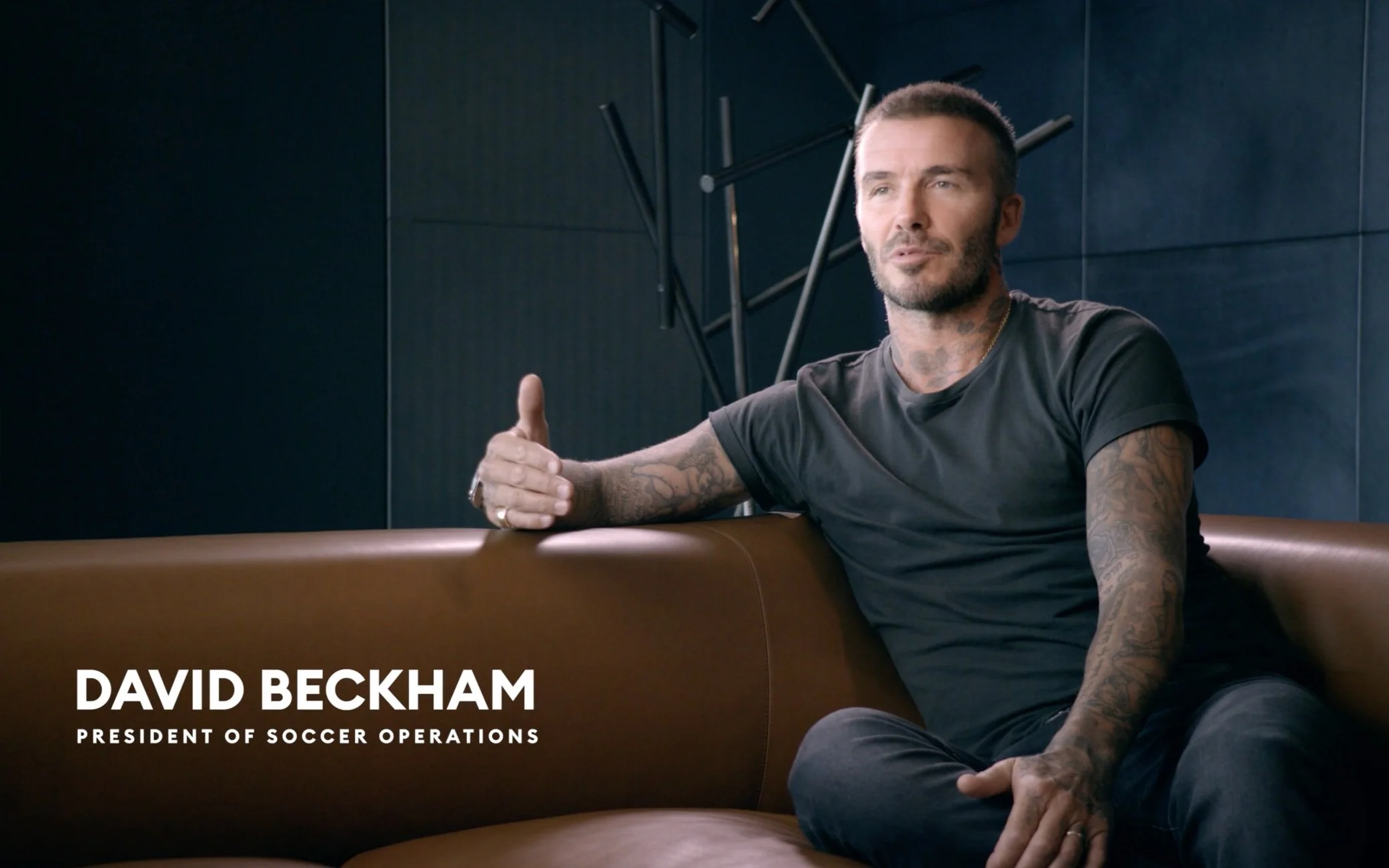 David Beckham sitting on a brown couch, wearing a black t-shirt, with a dark blue wall and modern art in the background. Text overlay: 'David Beckham, President of Soccer Operations'.