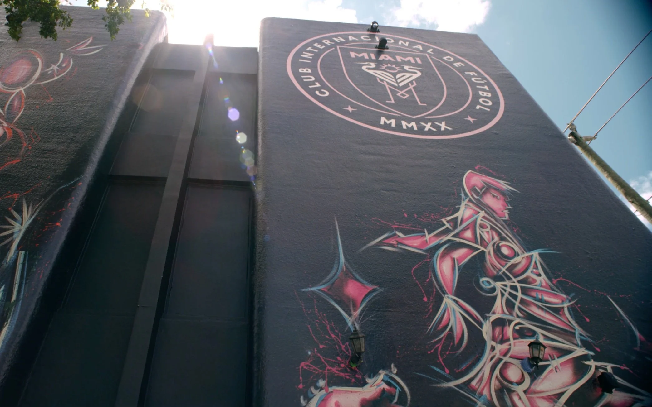 A tall building wall with a Miami Football Club mural and logo painted on it, featuring vibrant colors and graffiti-style art of a female character in pink and black tones. Sunlight creates lens flare in the upper left corner.