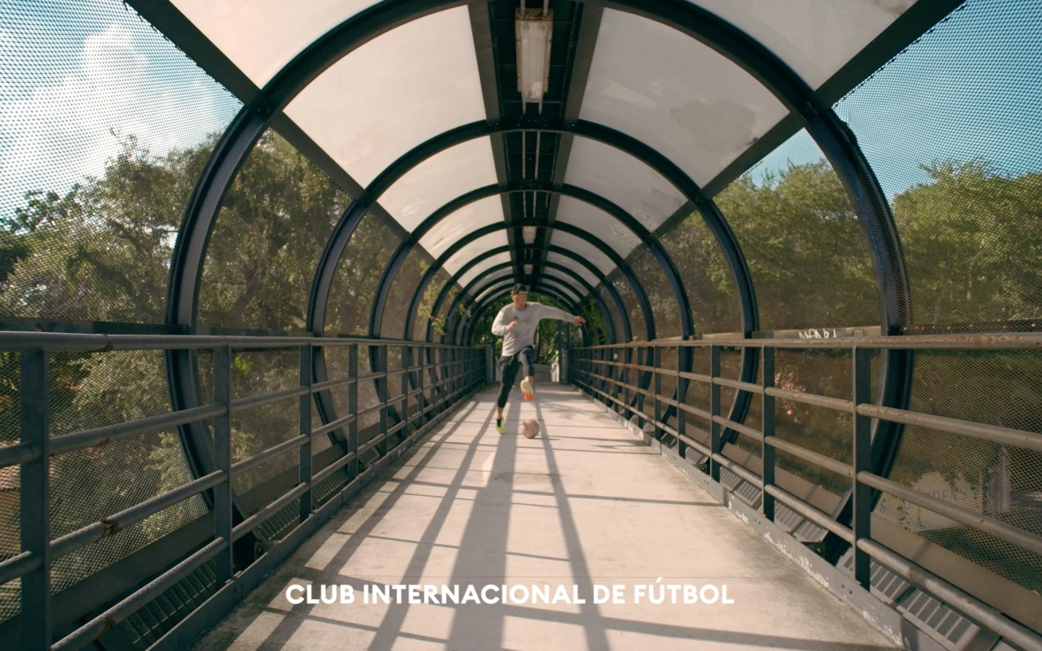 A person playing soccer on a footbridge with a curved, transparent roof and metal railings, surrounded by trees.