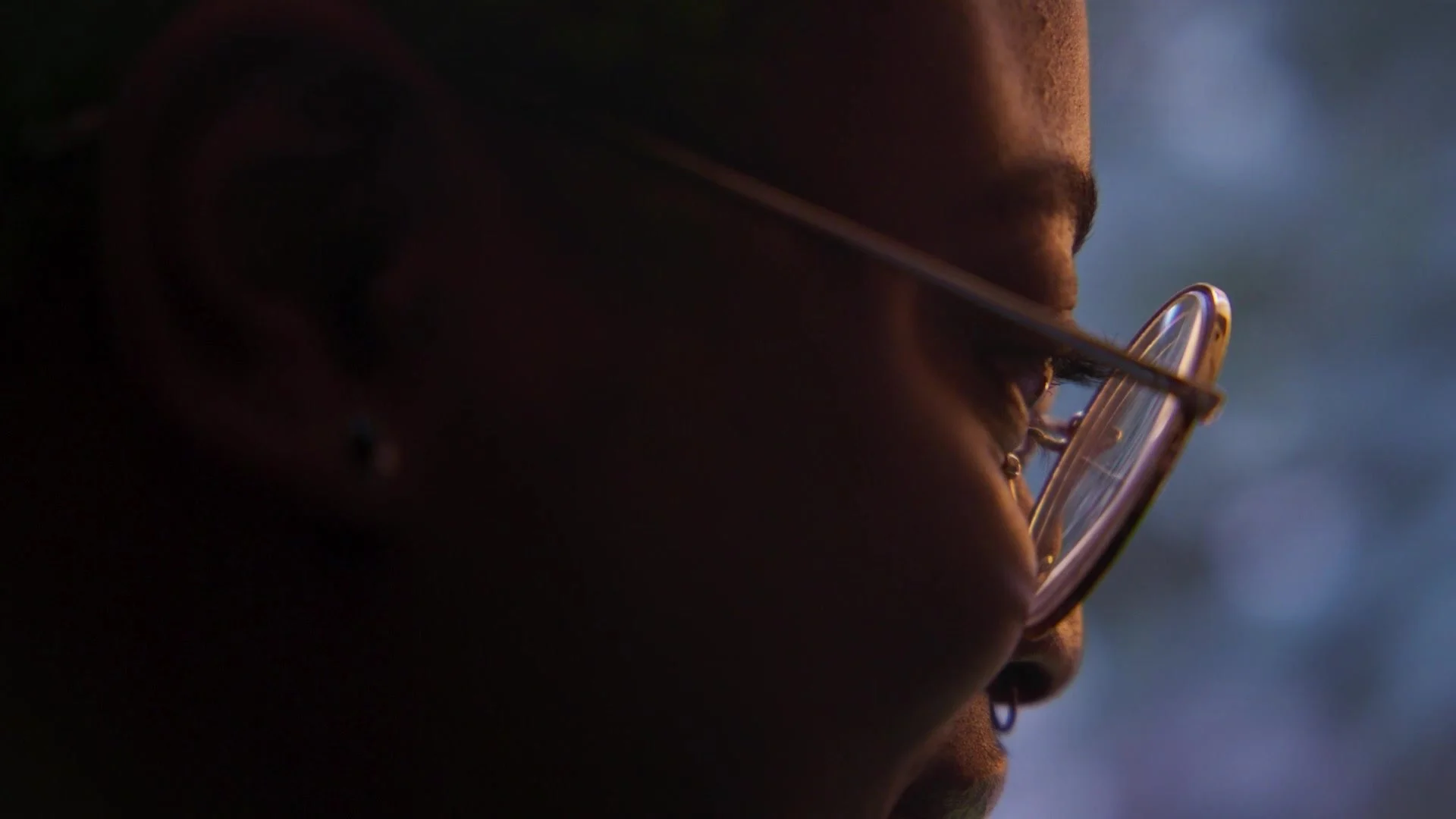 Close-up side profile of a person wearing glasses, illuminated by soft light, with a blurred background.