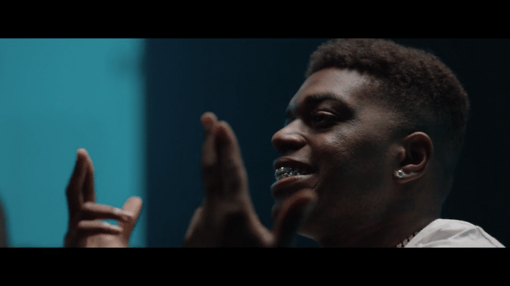 Close-up of a young man smiling and making hand gestures in a dark room with teal and dark background.