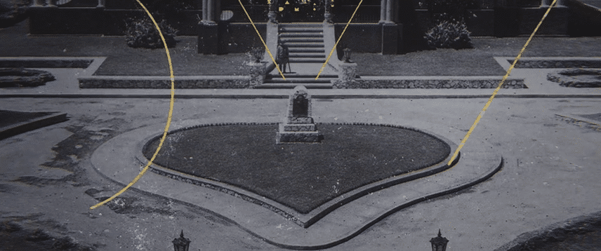 Nighttime scene of a small park with a heart-shaped centerpiece, surrounded by pathways and lamps, with a monument and stairs in the background.