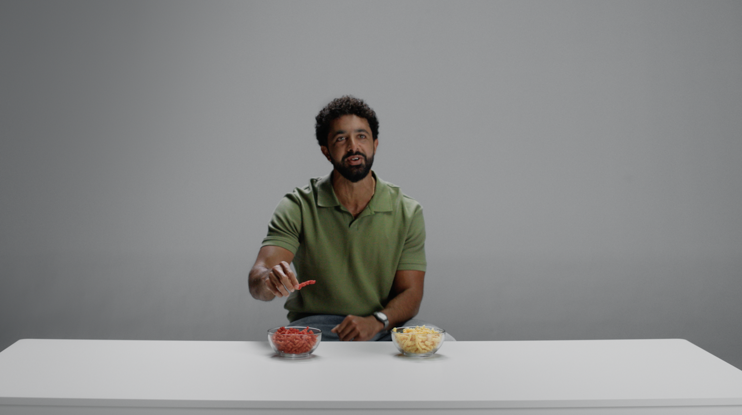 A man with a beard and curly hair sitting at a white table with two bowls of shredded cheese and diced tomatoes in front of him, against a plain gray background.