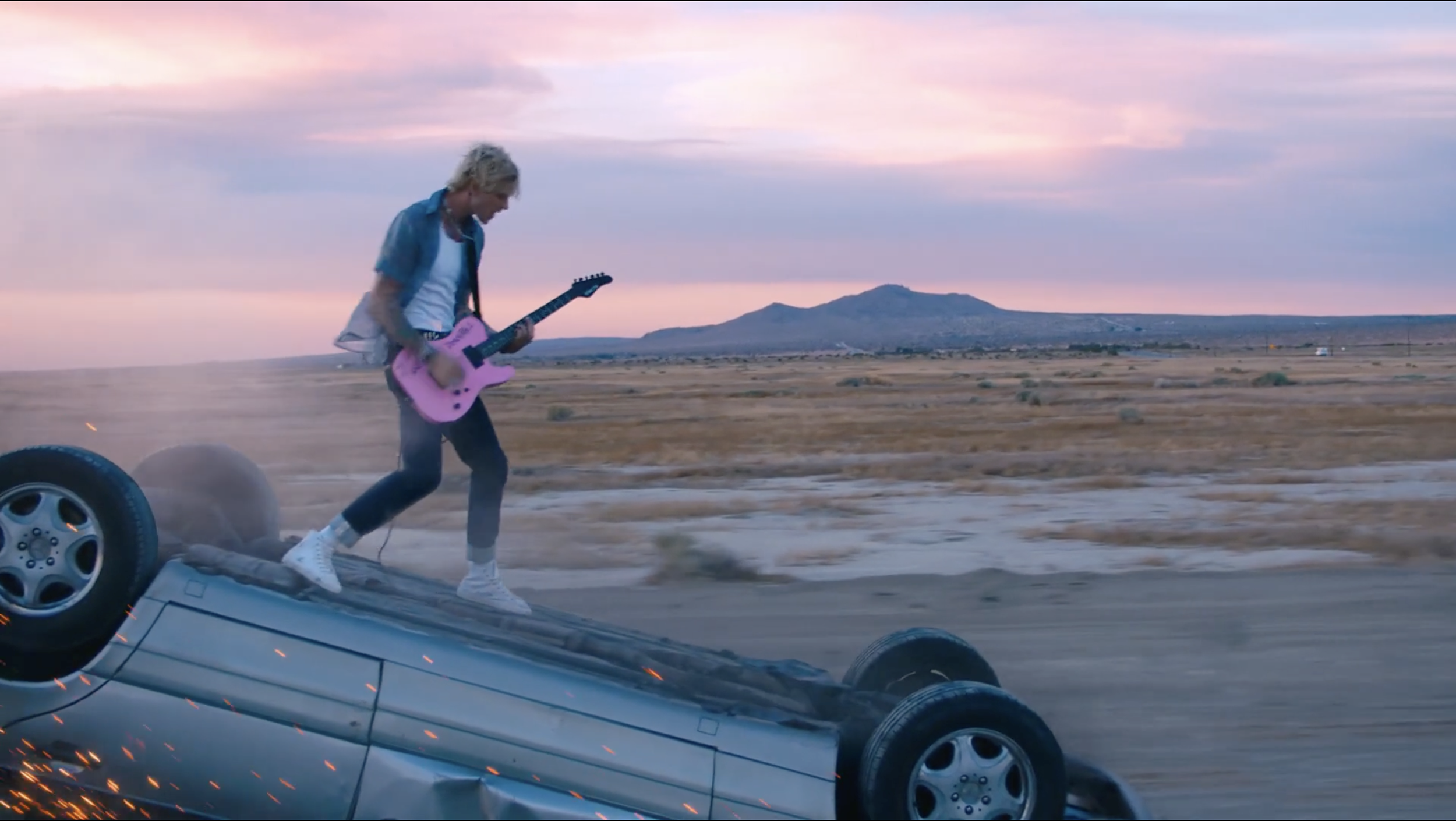 Person playing pink electric guitar standing on upside-down silver car in a desert at sunset.