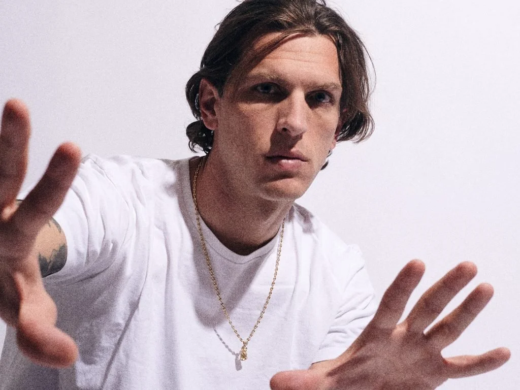 Young man with shoulder-length hair, wearing a white t-shirt and gold necklace, making a peace sign with both hands against a plain background.