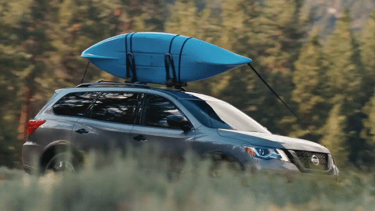A gray SUV with a blue kayak secured on top, parked outdoors with trees in the background.