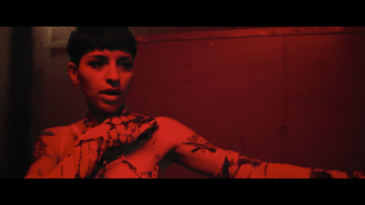 A woman with short dark hair and tattoos, illuminated by red lighting, looking toward the camera with a serious expression, against a dark background.