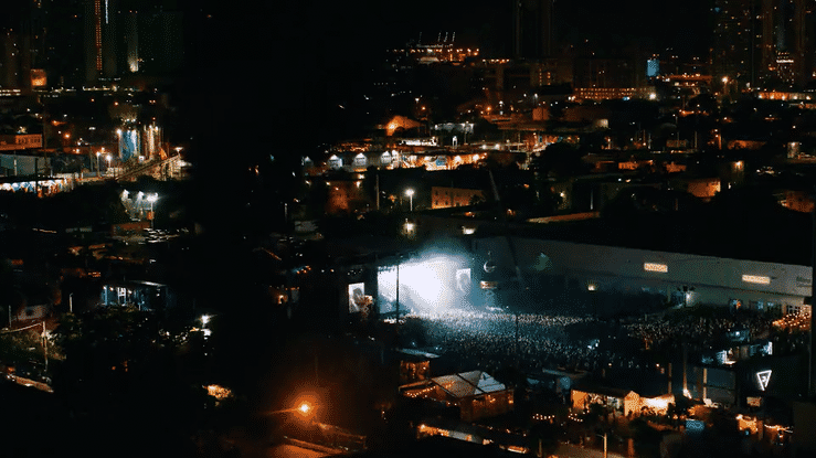Nighttime cityscape with brightly lit buildings, a lit-up outdoor concert or event space, and a dark sky.