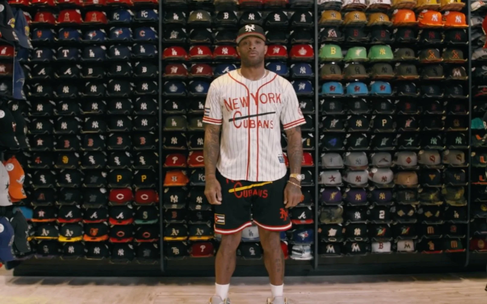 A man in a New York Cubans baseball uniform standing in front of a wall of various baseball hats in a store.