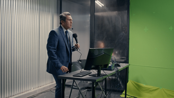 Man in suit presenting at a desk with computer and microphone in a conference room with green and gray walls.