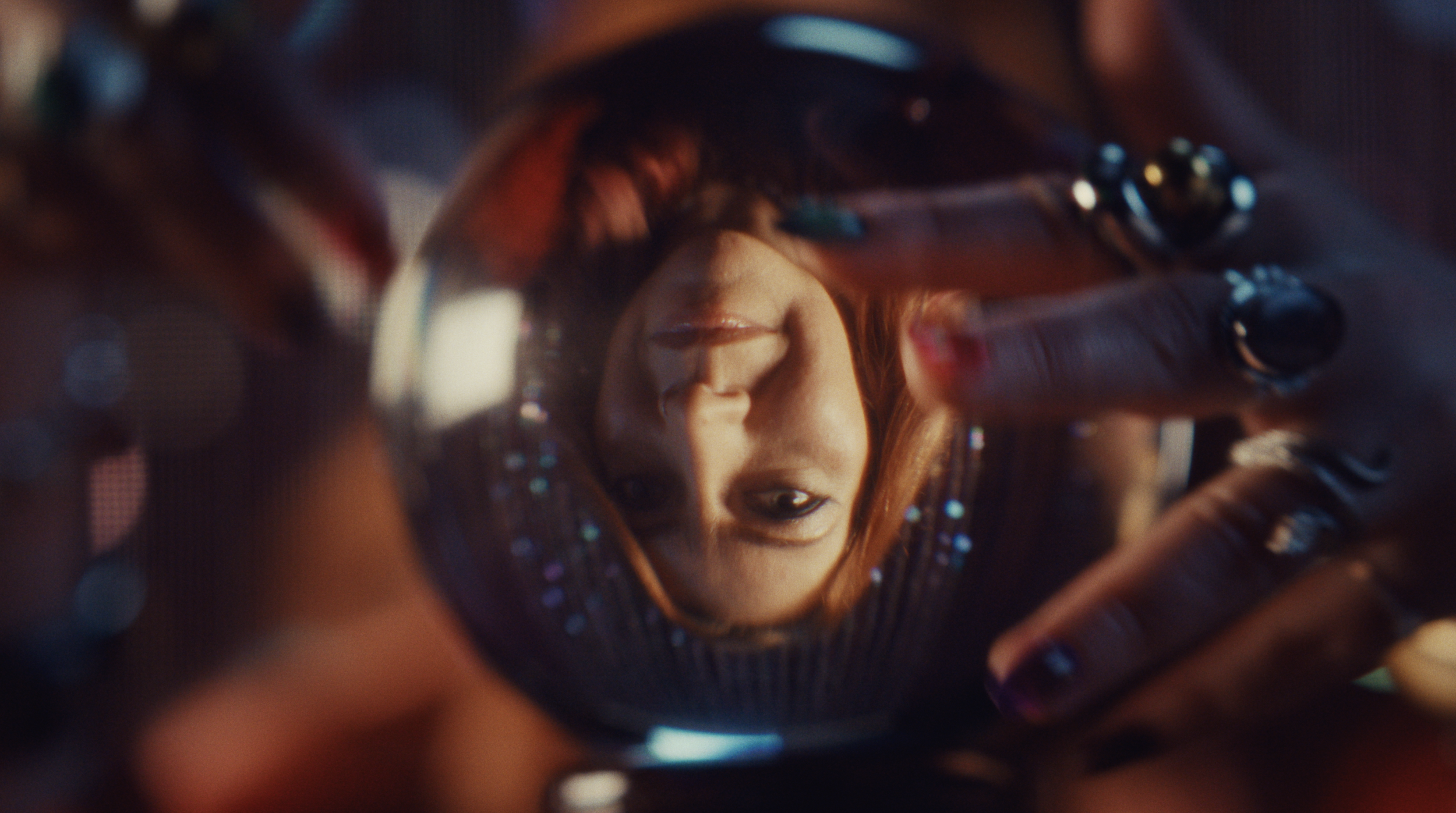 A woman with red hair and makeup looking into a camera through a reflective, circular metallic object, creating a distorted reflective effect.