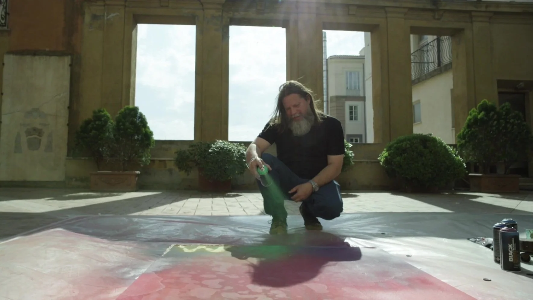 A man with long hair and a beard spray painting a large canvas on the floor inside an open-air building with potted plants in the background.