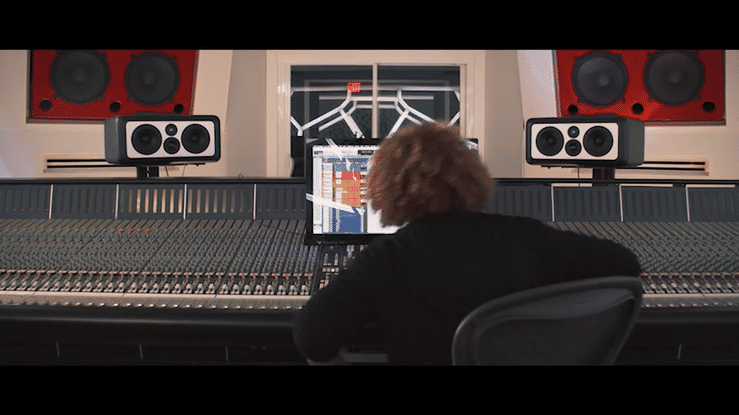 A person working at a large audio mixing console in a recording studio, with studio monitors and soundproof windows behind them.