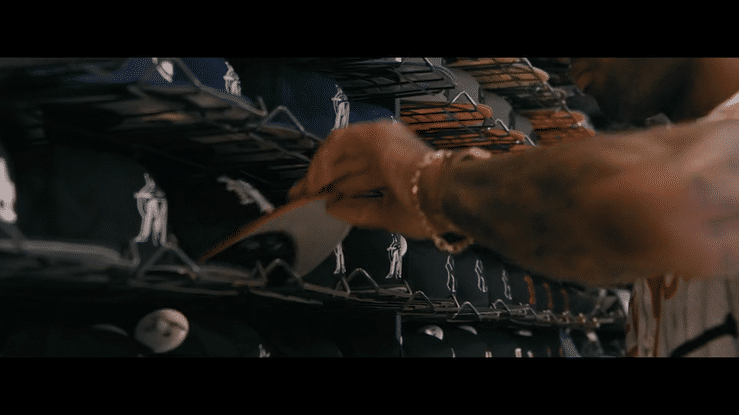 A person with tattoos and a watch is reaching for a black baseball cap with a white logo on a store shelf, which holds various caps.