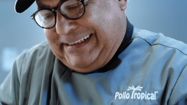 A man wearing glasses and a grey shirt with a Puerto Rico flag and the words 'Pollo Tropical' on it, smiling.