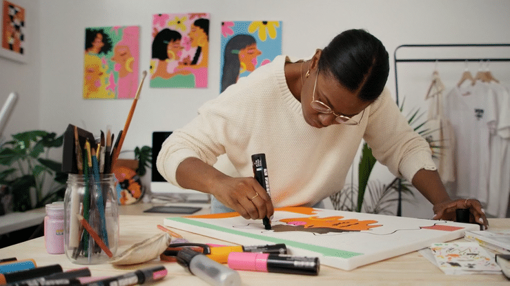 Woman working on colorful artwork in an art studio with paintings on the wall and art supplies on the table.
