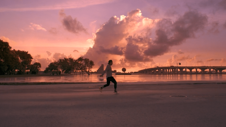 Person running along a beach during sunset with clouds, trees, water, and a bridge in the background.