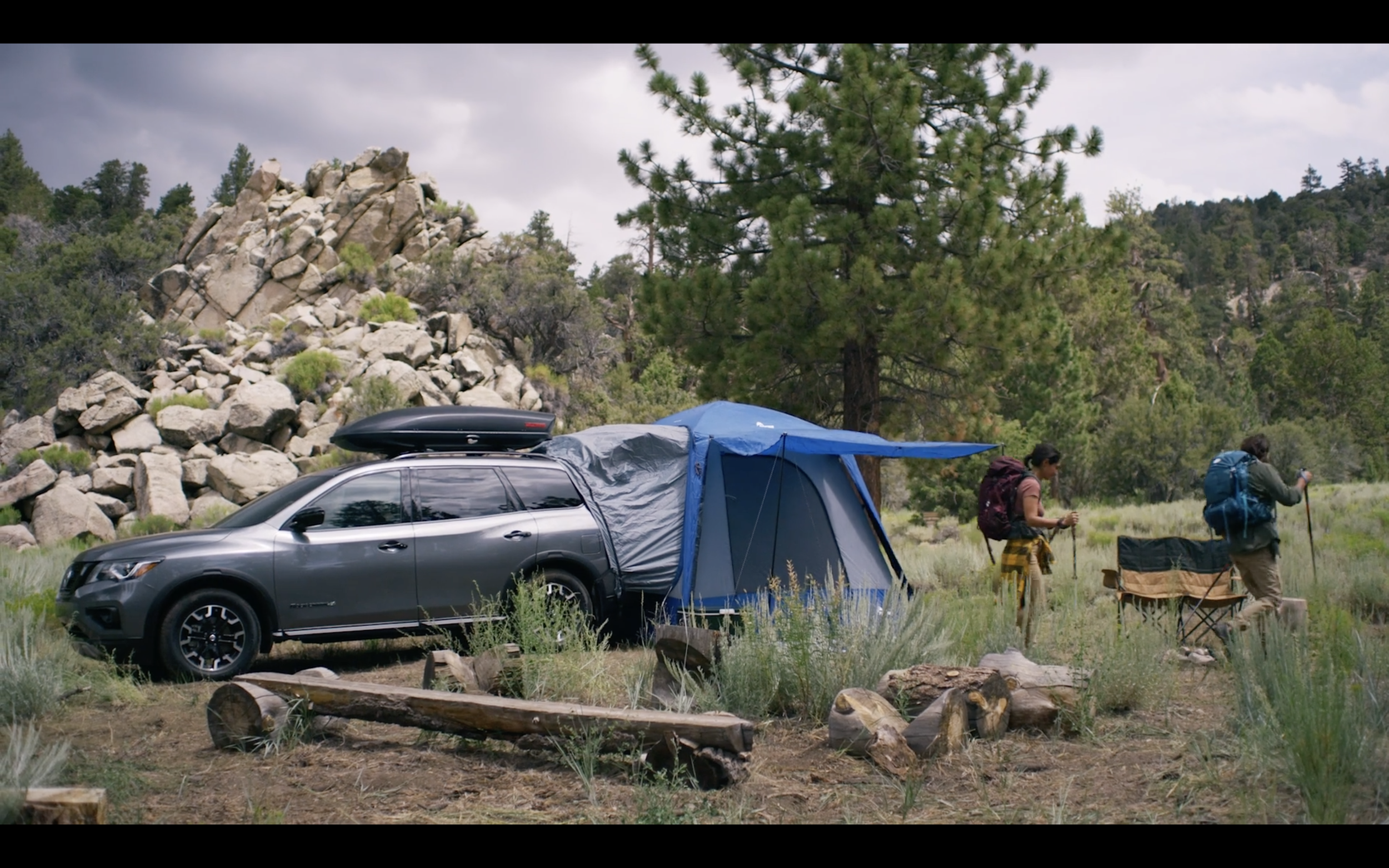 Two people camping near a black SUV with a blue tent attached, in a forested area with trees, rocks, and logs.
