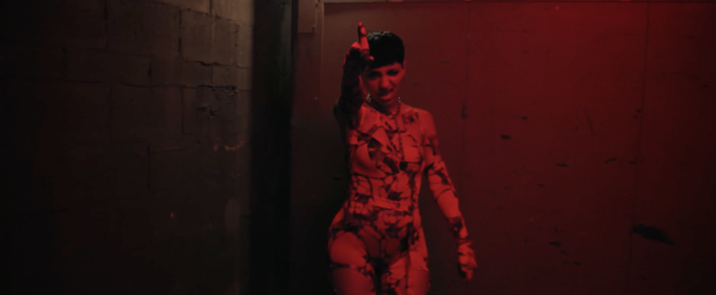 A woman standing in a dimly lit room with red lighting, wearing a patterned dress and pointing upward.