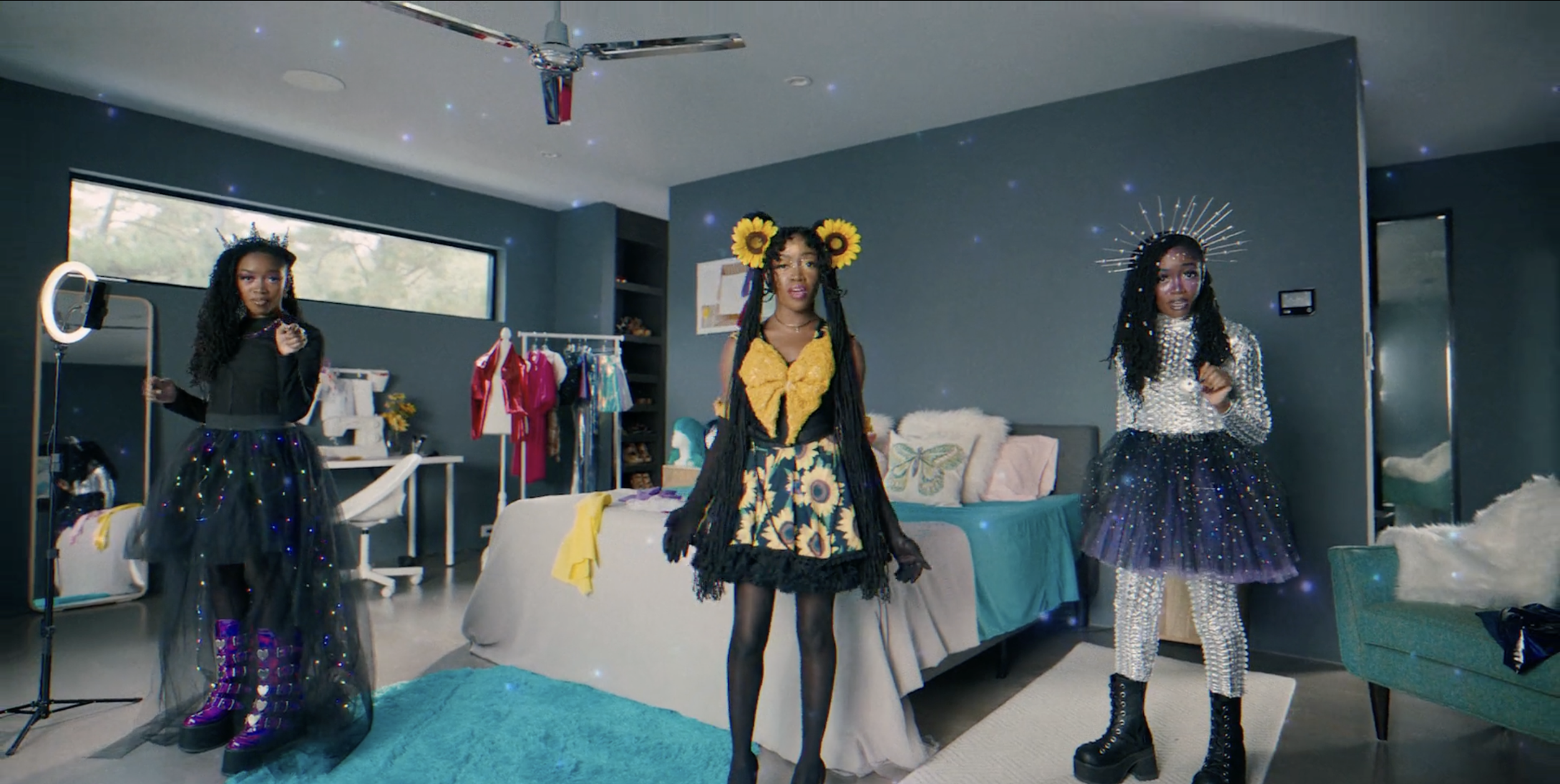 Three women dressed in fashionable, glittery, and colorful outfits standing in a modern bedroom with artistic decor and glowing starry overlay.