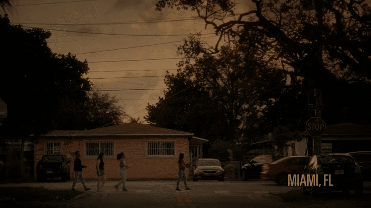 A group of people walking across a street in Miami, Florida, during sunset or dusk, with trees and parked cars visible along the street.