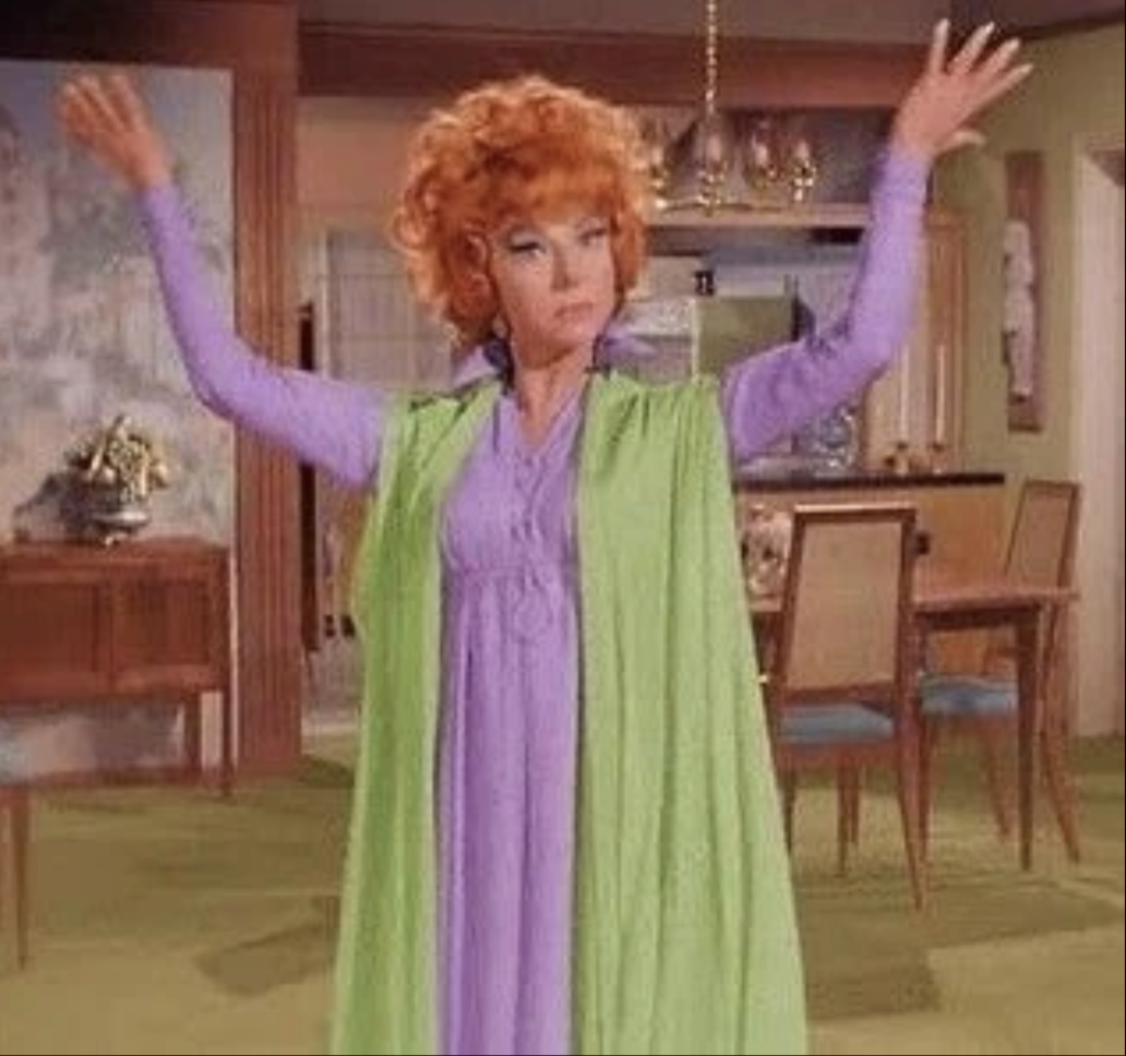 A woman with curly red hair standing in a dining room with her arms raised, wearing a lavender dress and a green cape.