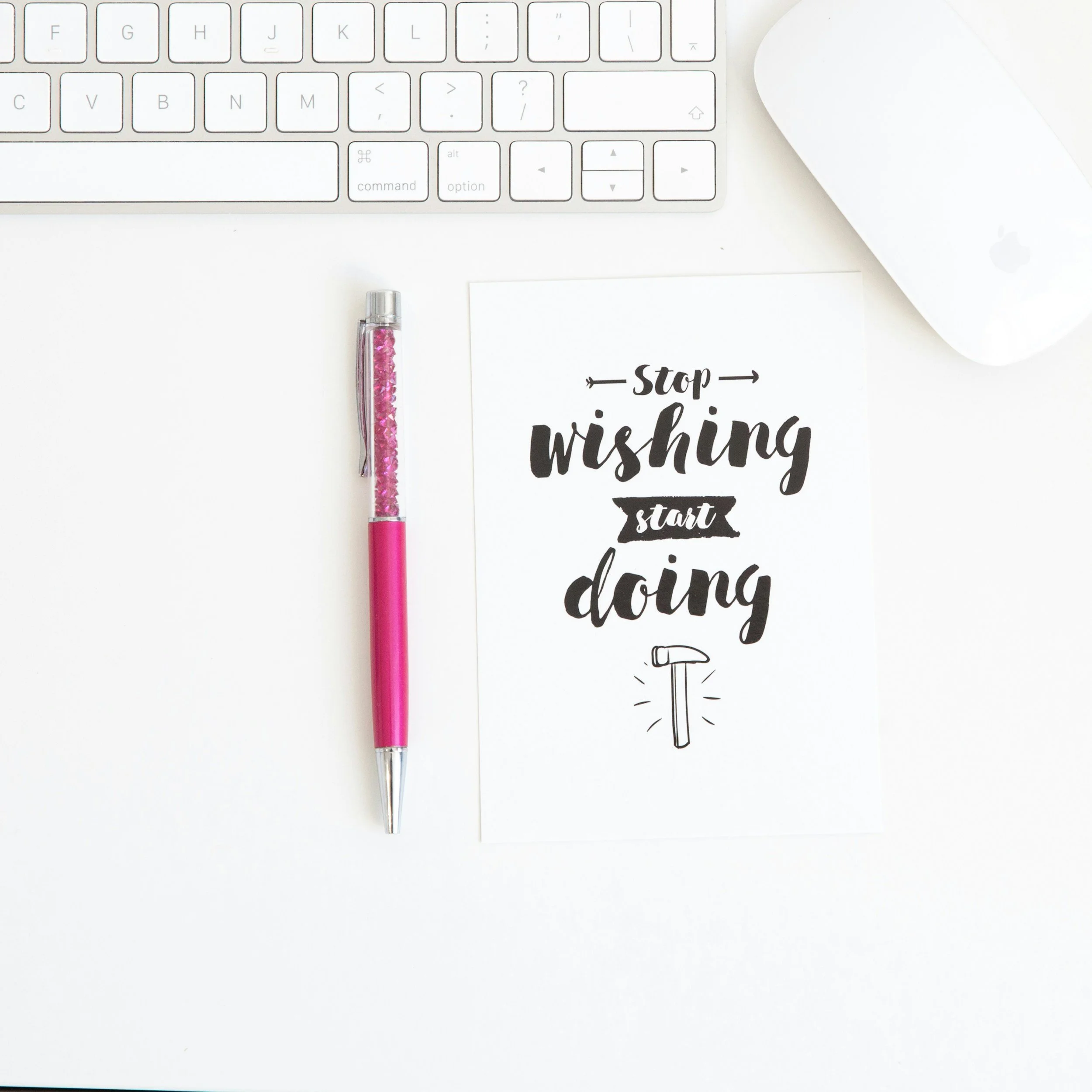 A pink and silver pen, a motivational quote on paper that says "Stop wishing start doing" with a hammer illustration, a white computer keyboard, and a white wireless mouse on a white desk.