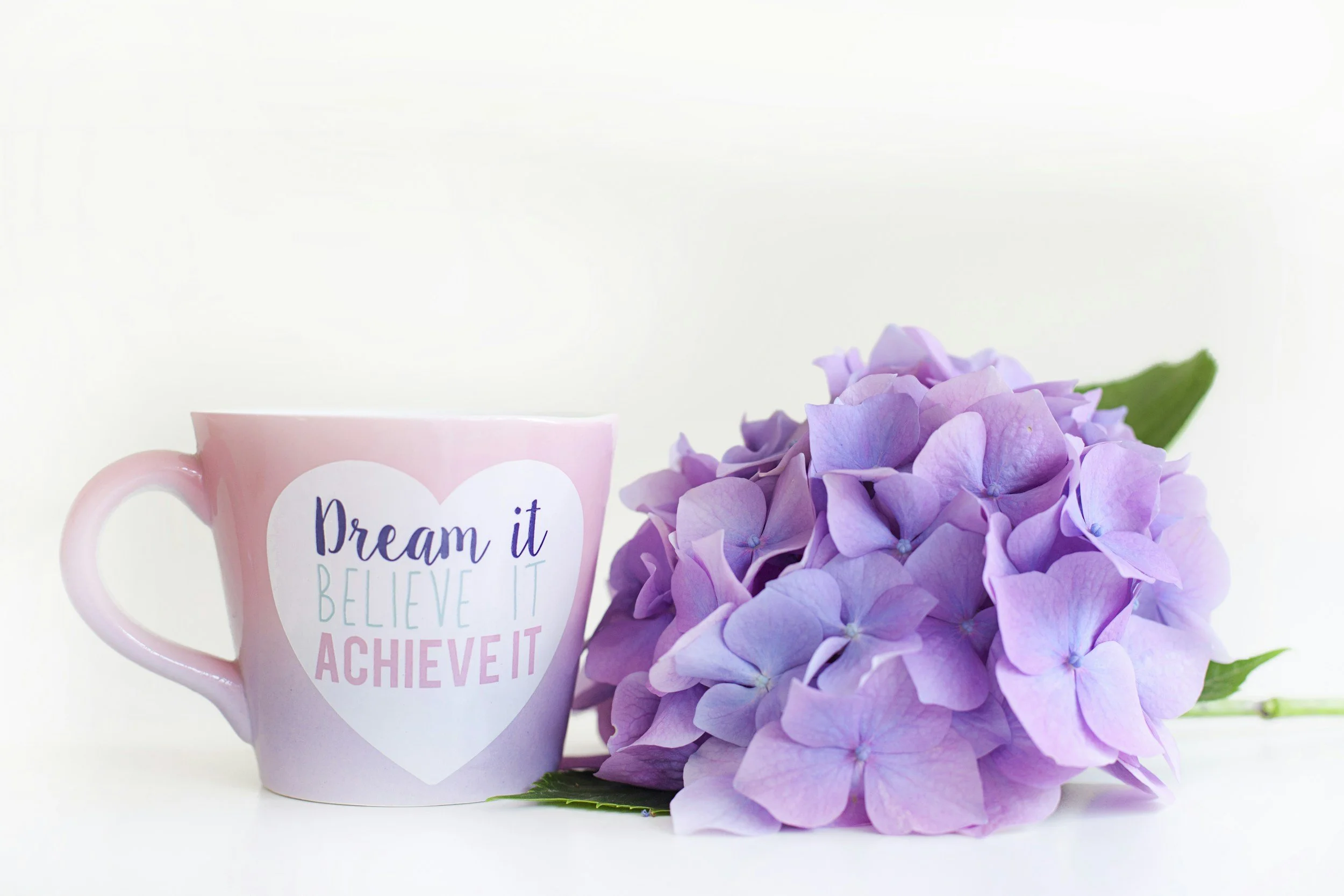 Pink mug with motivational quote next to a bunch of purple hydrangea flowers.