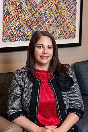Woman sitting on a beige couch in front of a colorful abstract painting, wearing a black and white jacket and red top.