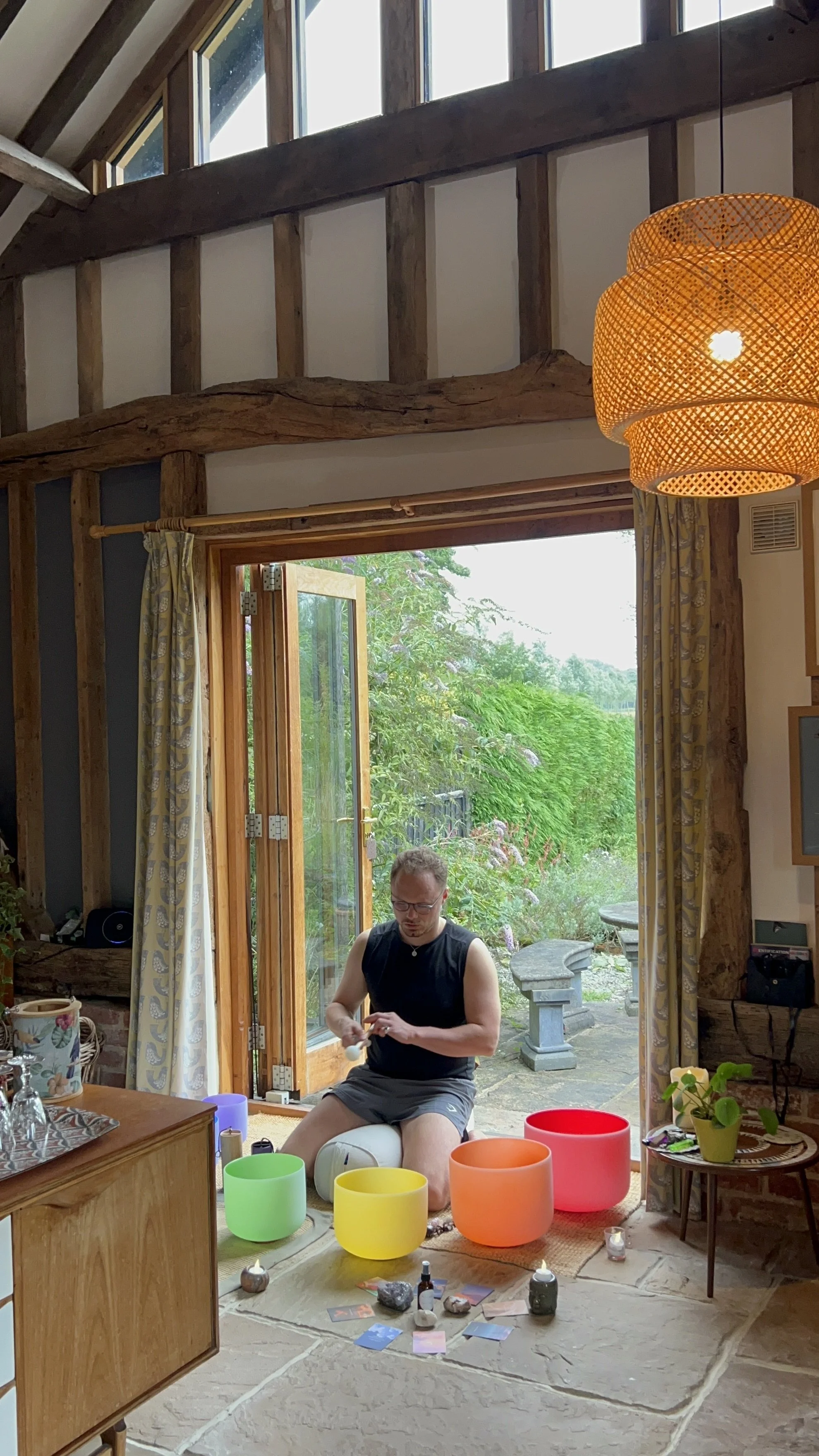 A man sitting on a stool playing colorful singing bowls in a cozy, rustic room with large open door to a garden, natural light, and wooden interior.