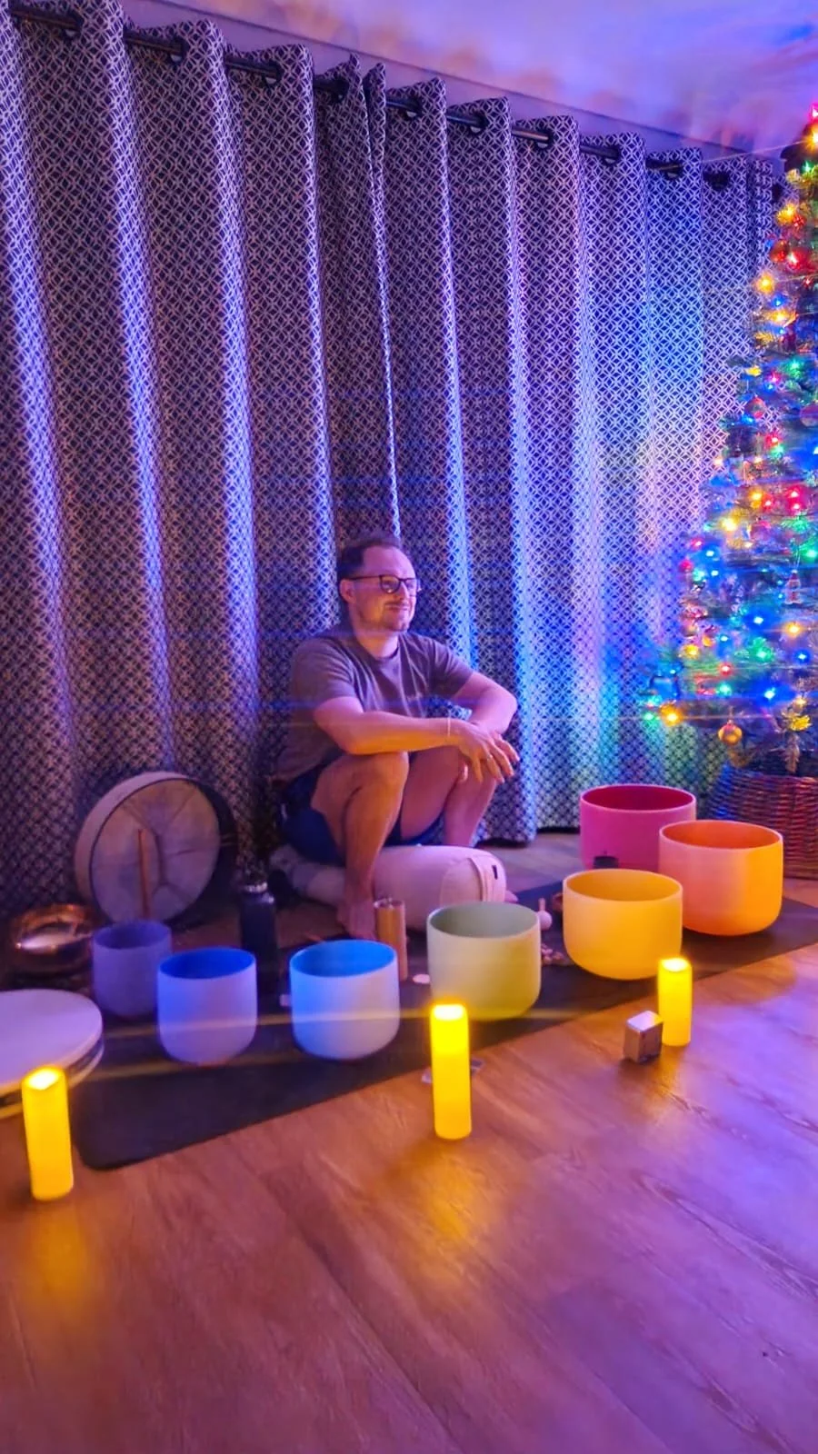 A person sitting on a meditation cushion in a room decorated with a Christmas tree and colorful singing bowls, illuminated by LED candles, with patterned curtains in the background.