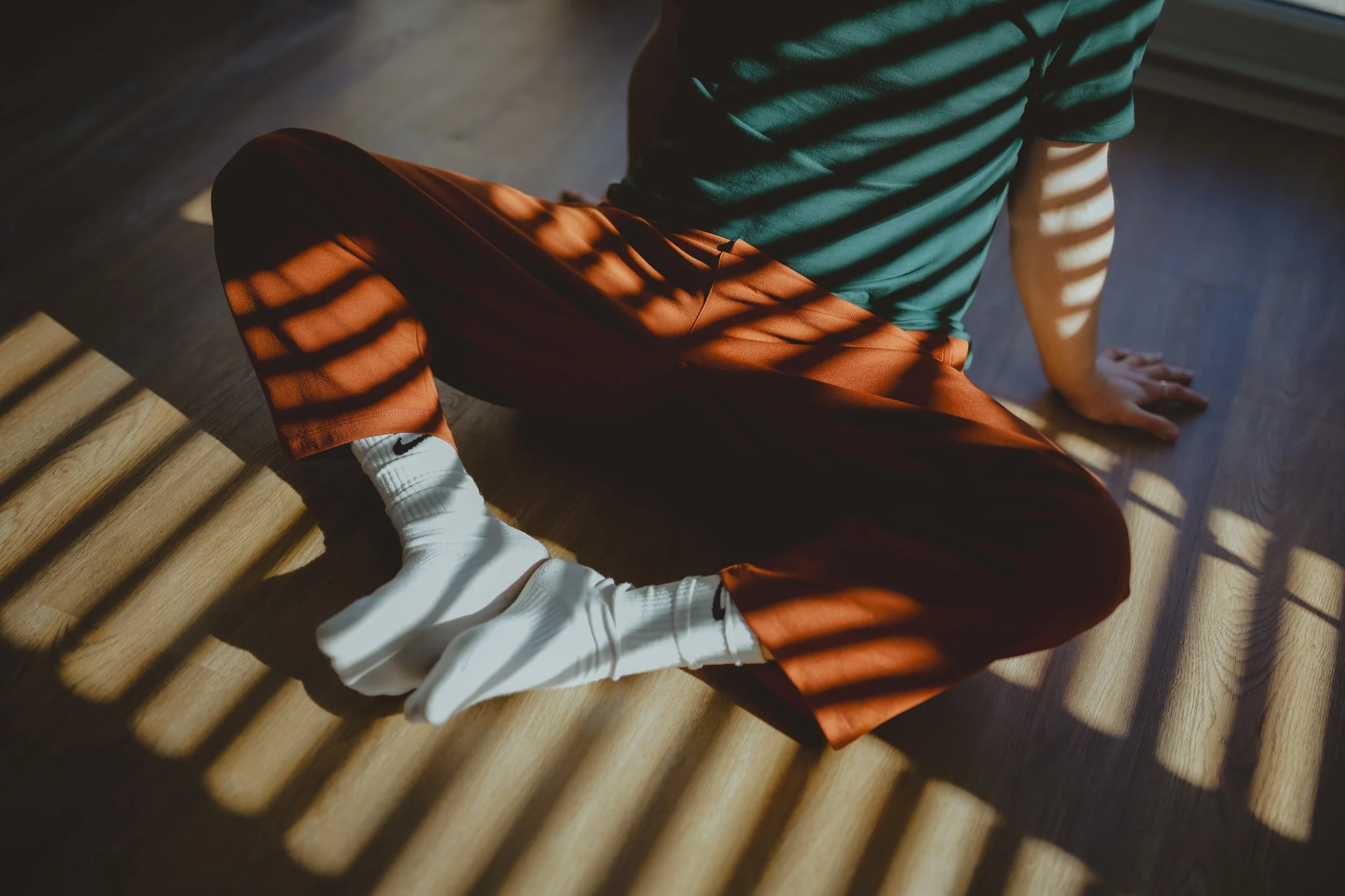 Person sitting on the floor with striped shadows cast by window blinds, wearing white socks, rust-colored pants, and a dark green shirt.