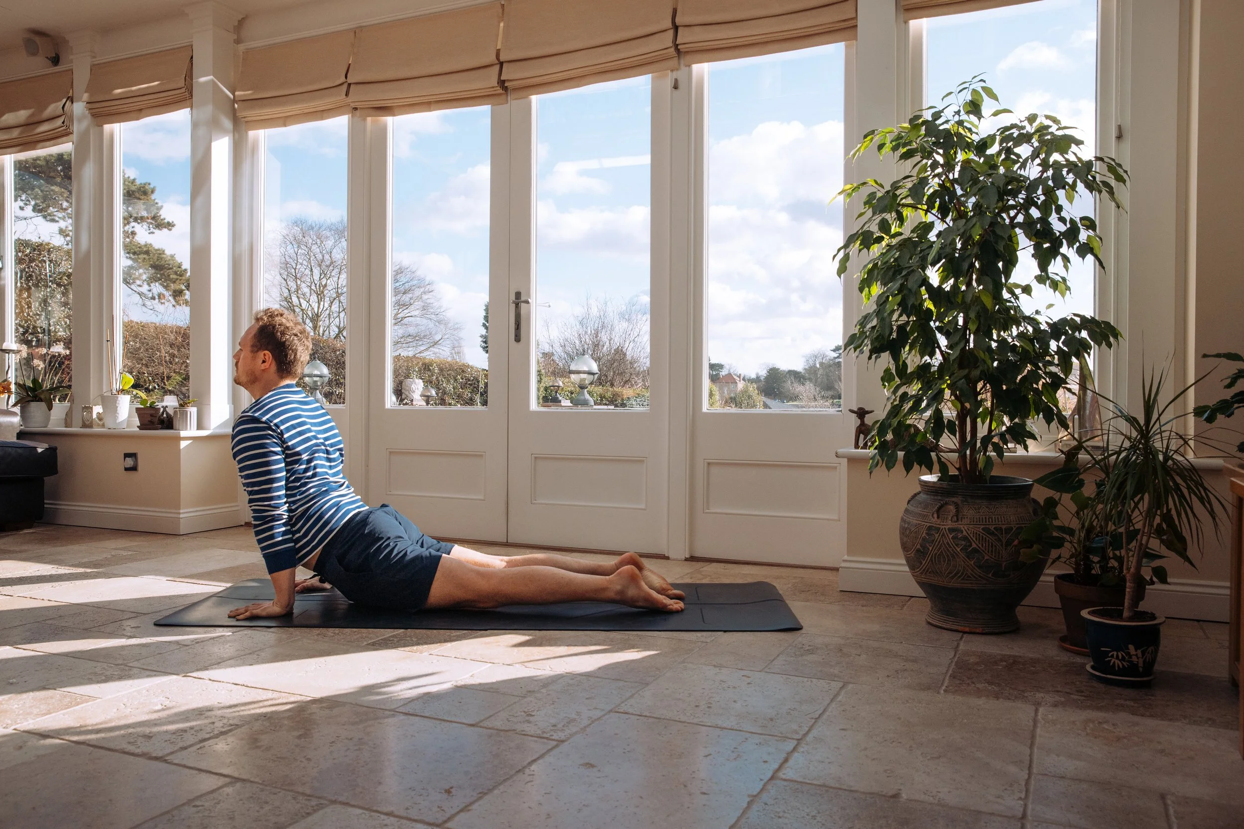 A man doing the cobra pose yoga in a sunlit room with large windows, potted plants, and tiled floors.