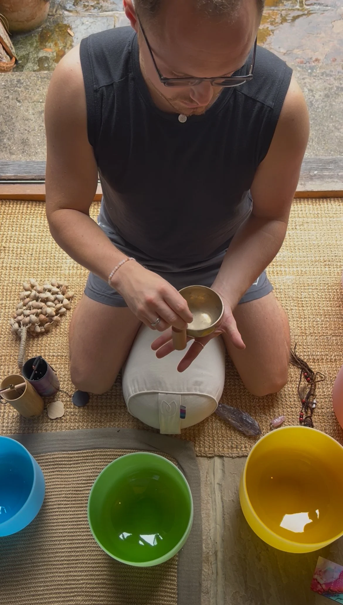 A man sitting on a stool playing colorful singing bowls in a cozy, rustic room with large open door to a garden, natural light, and wooden interior.