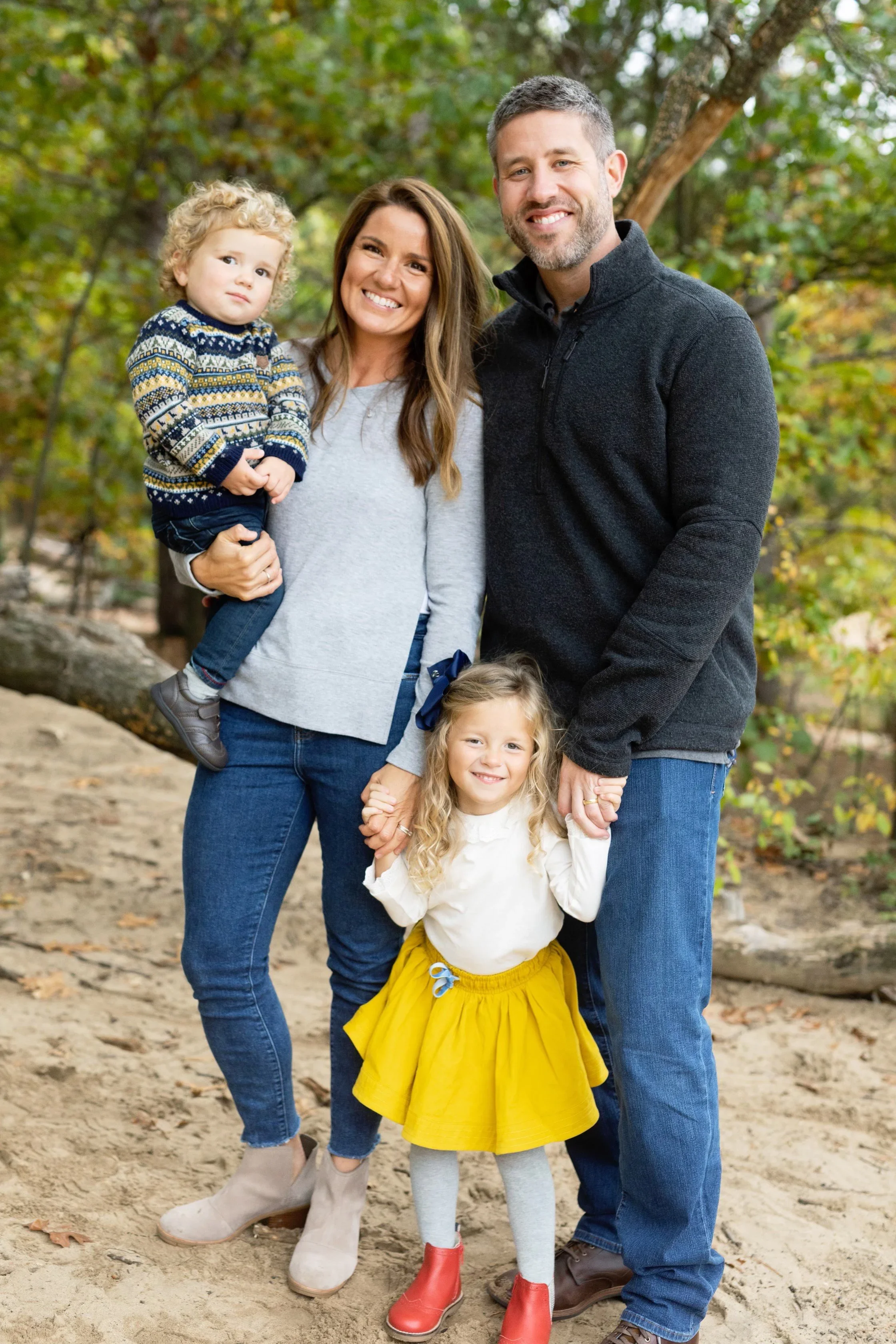 A happy family of four outdoors in a wooded area, with a woman and a man smiling, and two little girls, one in a yellow skirt and the other in dark pants, all holding hands or being held.