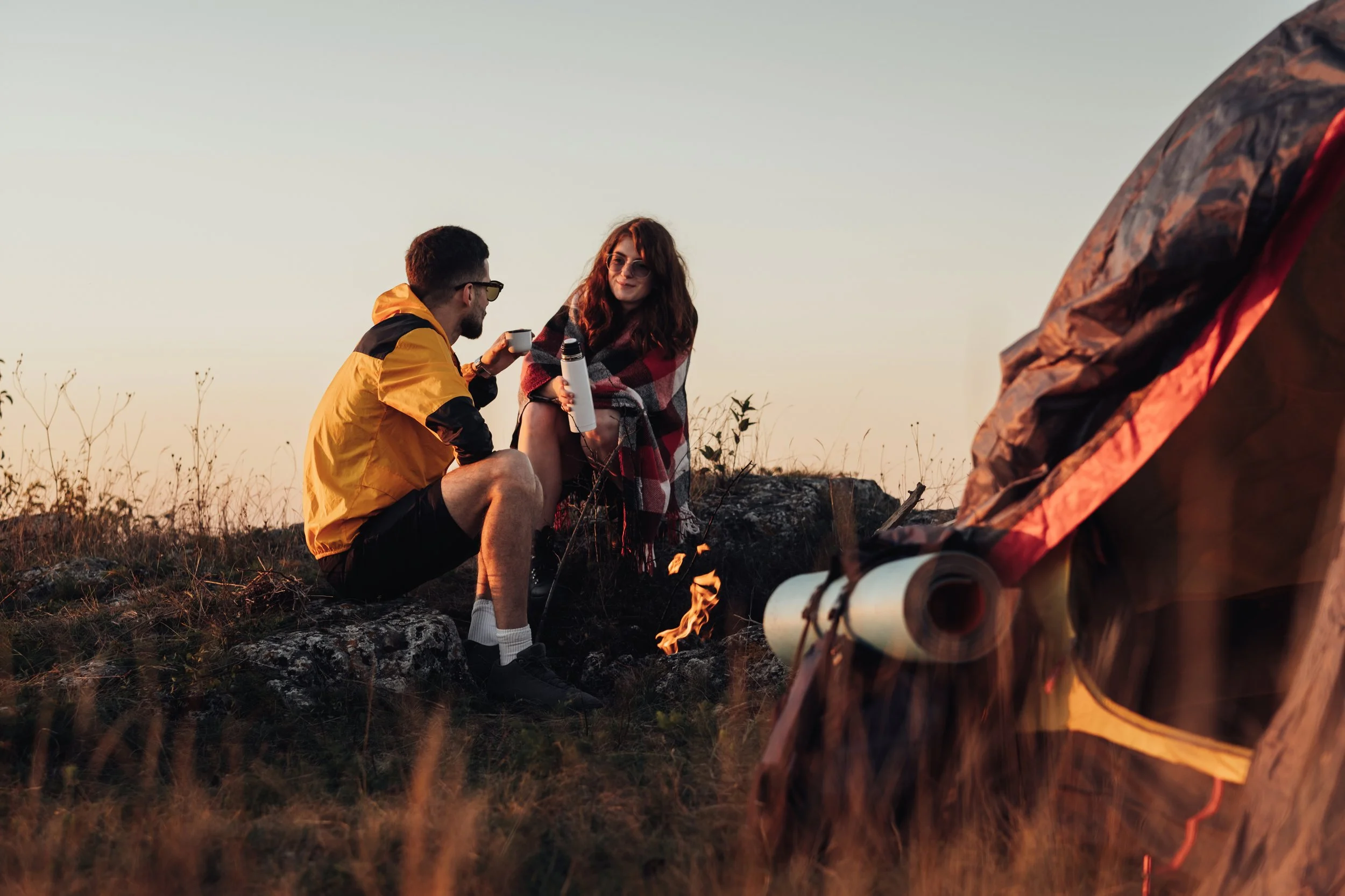 Two young adults sit around a campfire outdoors, sharing drinks, with camping gear visible, during sunset or late afternoon.