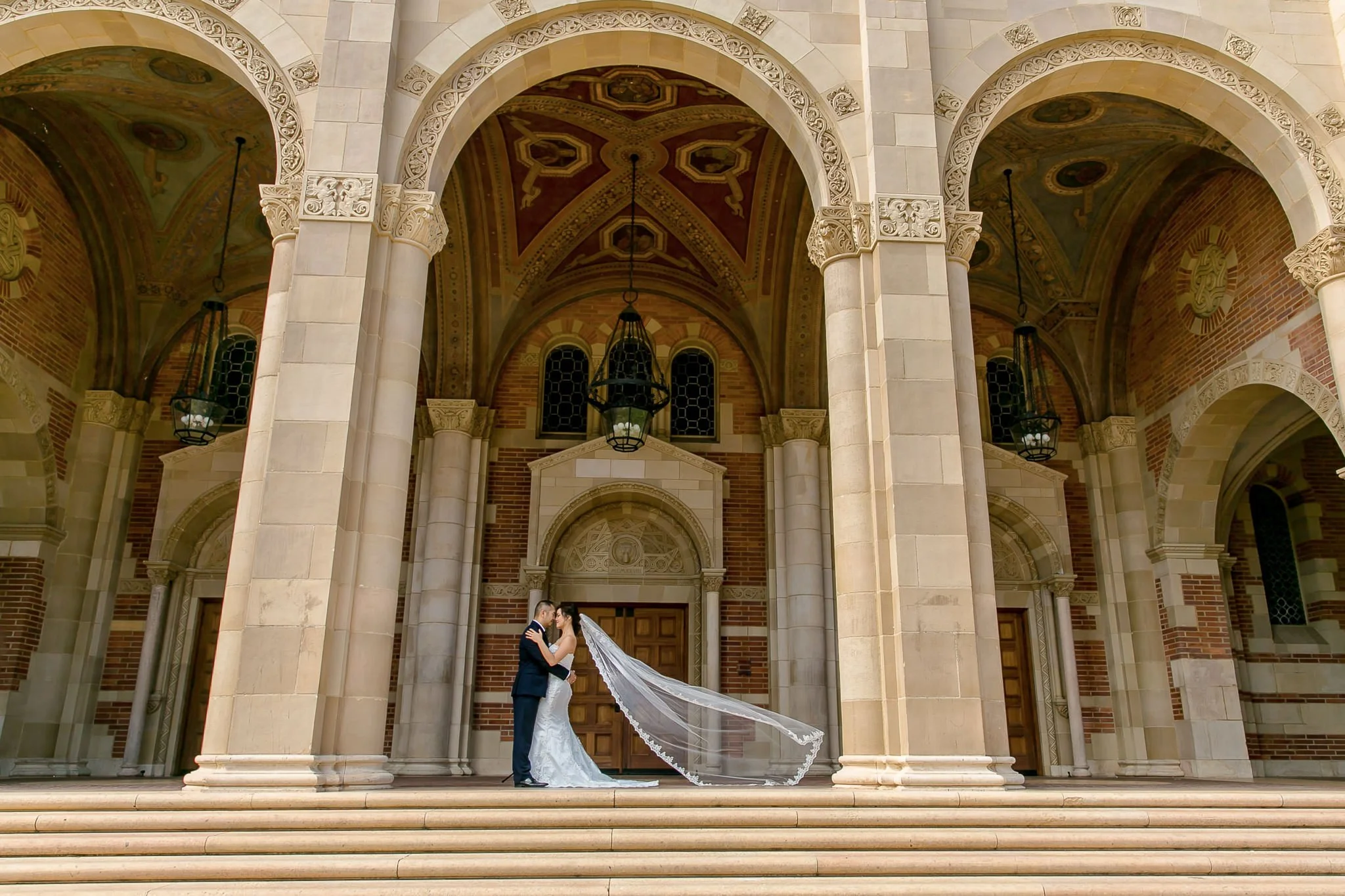 Bride and groom standing in a high-contrast editorial pose against a stone archway, with the groom's arm around bride and veil floating in the wind.