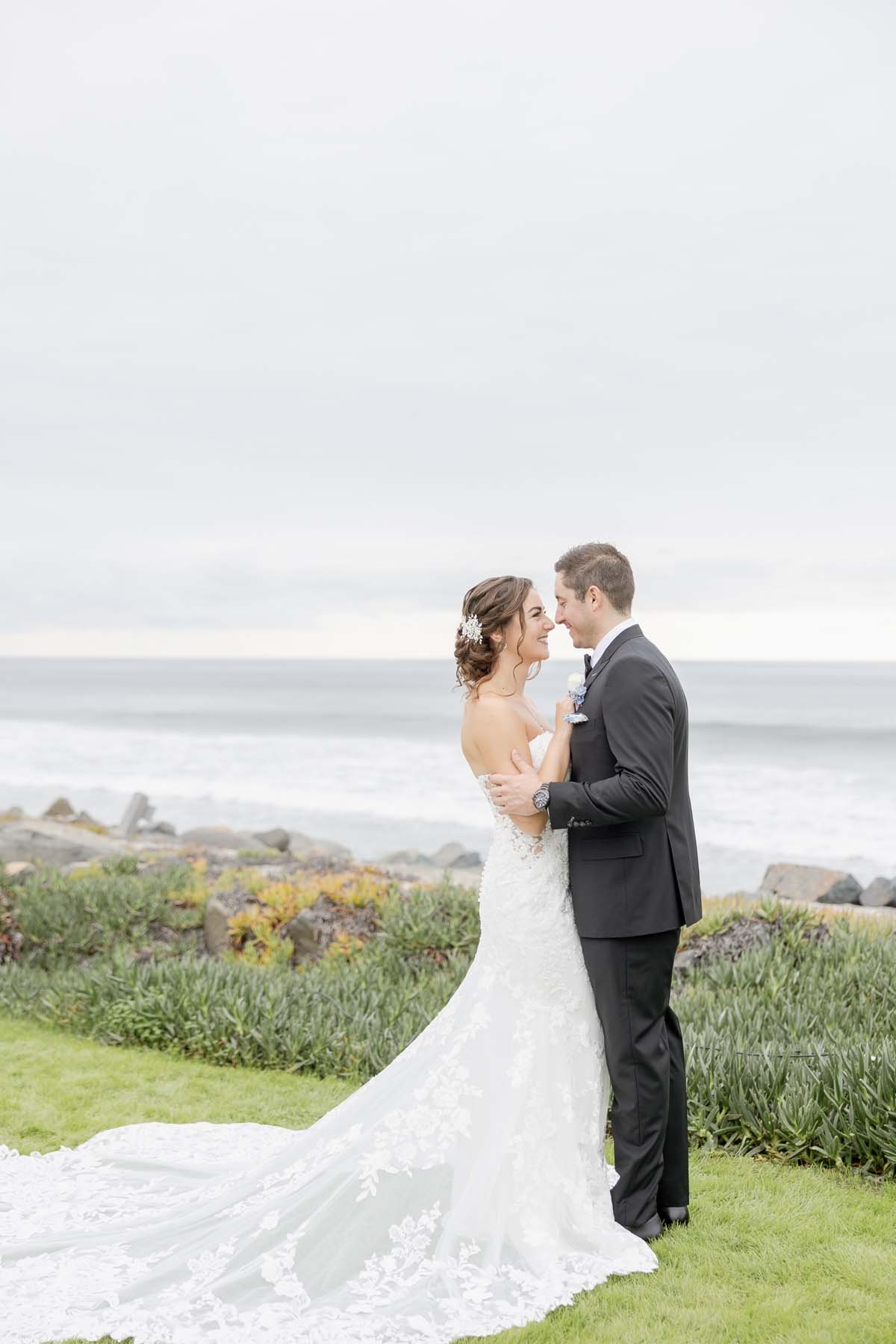 Bride and groom embracing outdoors by the ocean with greenery and rocks.