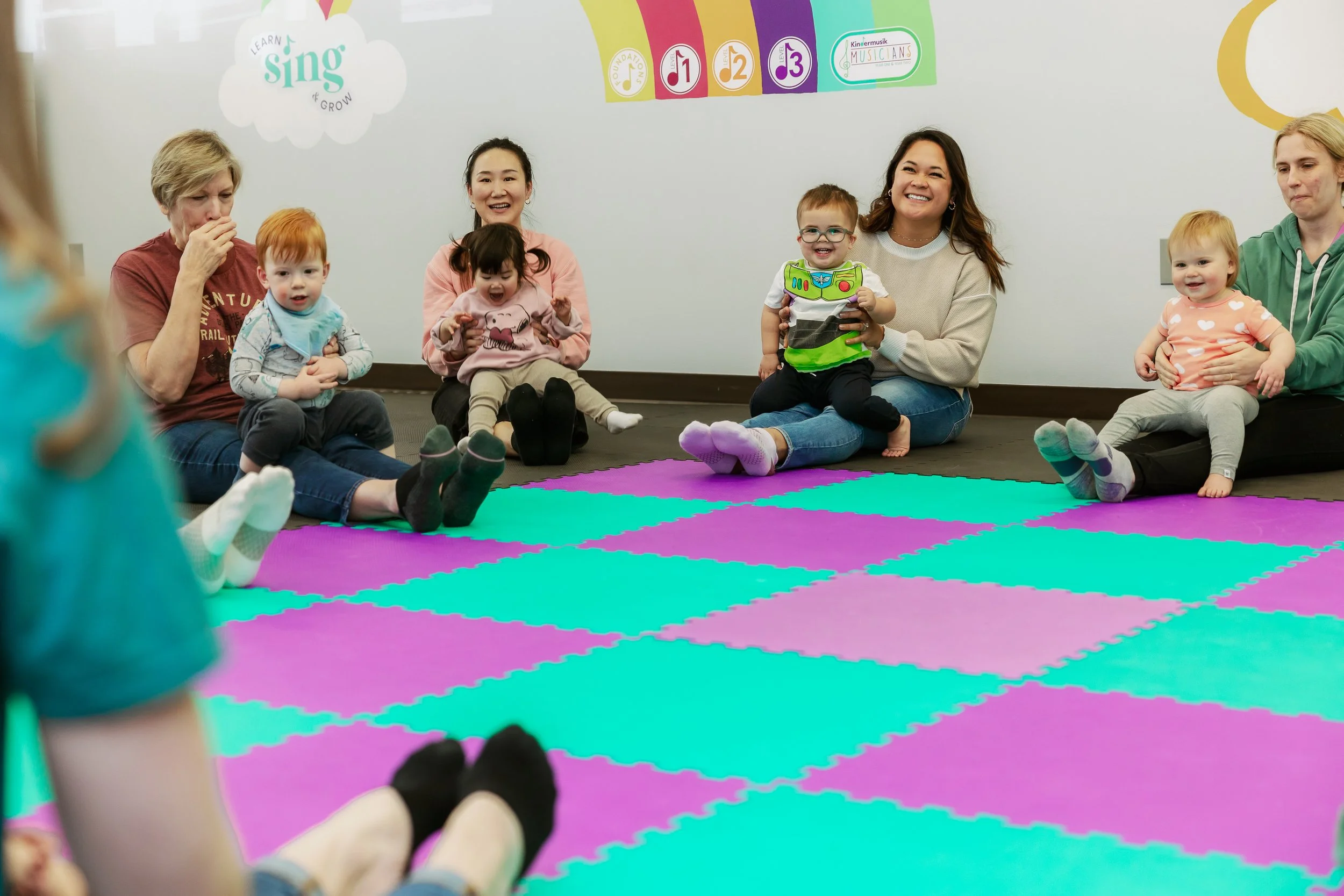 Children and adults sitting on a colorful foam mat in a classroom, engaging in a singing or music activity, with a white wall and colorful educational posters in the background.