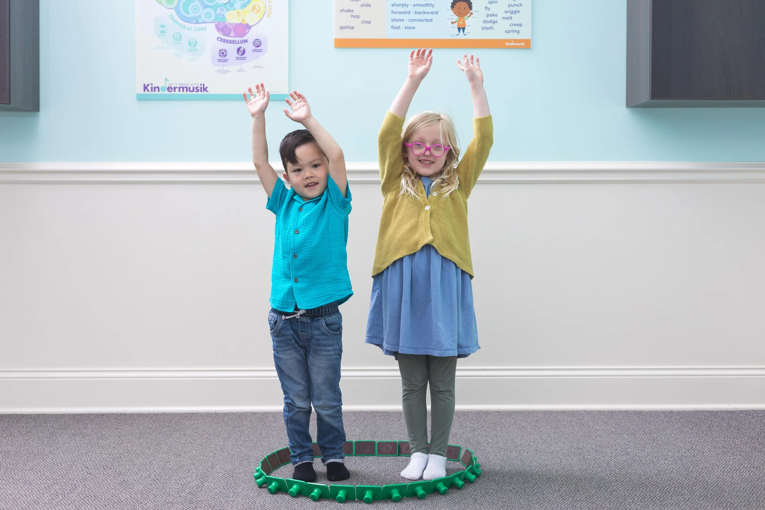 Two children, a boy and a girl, standing inside a green circular track in a room with educational posters on the wall. The boy is wearing a blue shirt and jeans, while the girl is wearing glasses, a yellow cardigan, and a blue dress. Both children have their arms raised above their heads.