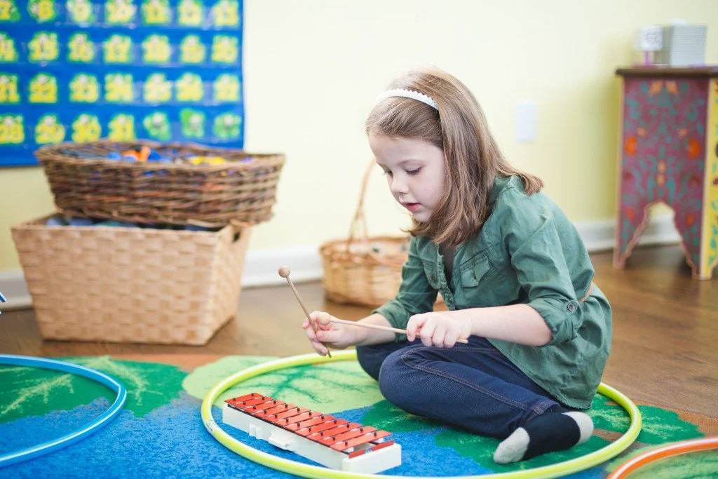 A young girl sitting on a colorful rug, playing a xylophone with mallets, in a classroom with baskets and wall decorations.