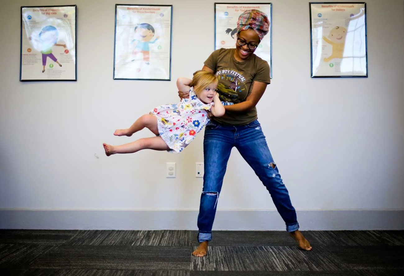 A woman with a headwrap and glasses holding a young girl with red hair in a floral dress, lifting her up playfully in a room with framed posters on the wall.