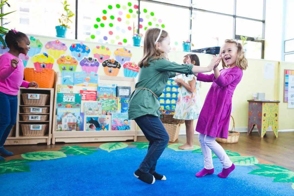 Two girls playing and laughing together on a blue rug with green leaf patterns in a colorful classroom. One girl in a purple dress is holding hands with another girl in a green top and jeans, who is also smiling. Other children are in the background, with classroom decorations including books, pictures of cupcakes, and colorful balloons.