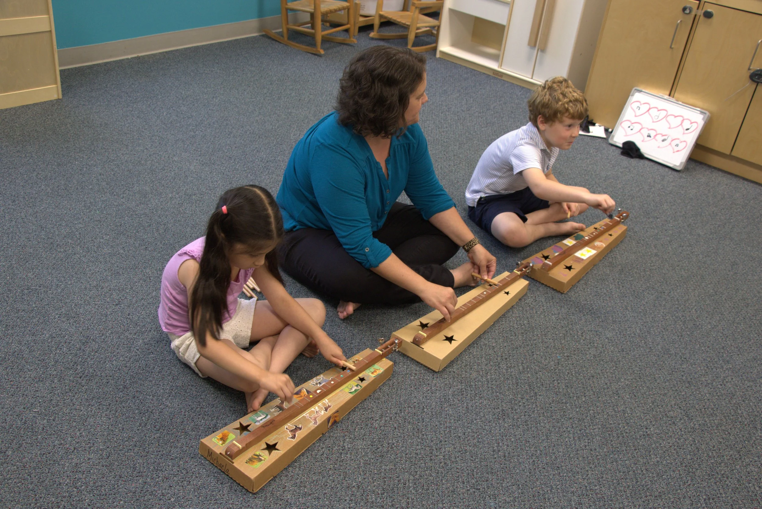 A woman and two children sitting on a carpeted floor playing with a long wooden toy with animals and shapes. The woman has short brown hair and is wearing a blue shirt. The boy has curly red hair and is wearing a striped shirt. The girl has long dark hair with a pink hair tie and is wearing a pink shirt. In the background, there is a whiteboard with colorful hearts and a collection of wooden chairs and shelves.