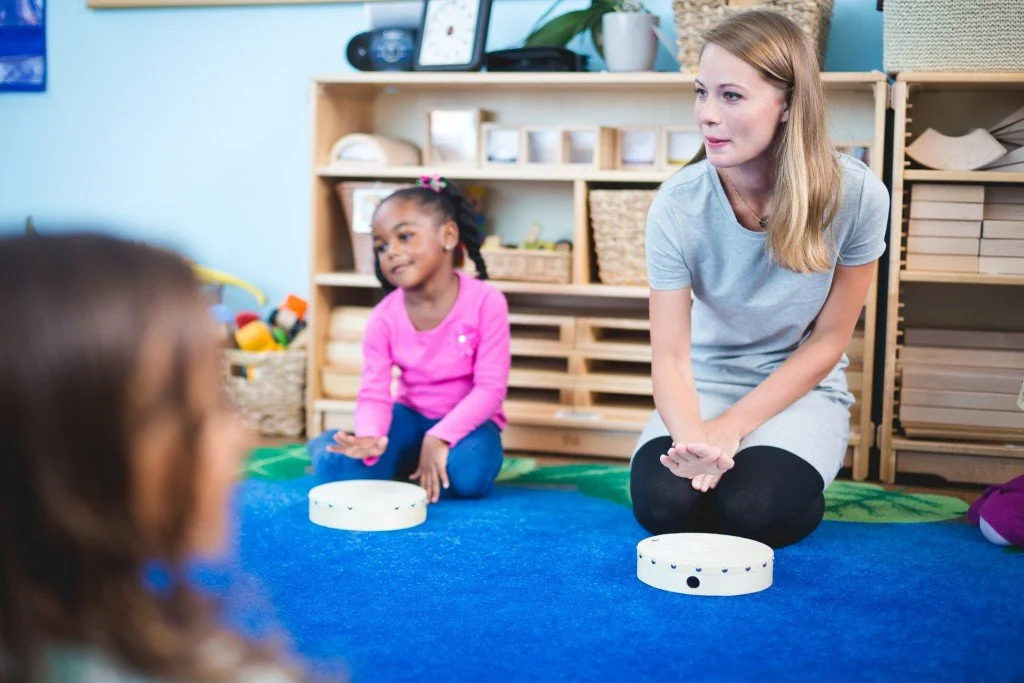 A woman and two young girls sitting on a blue carpet in a classroom, engaged in a group activity.