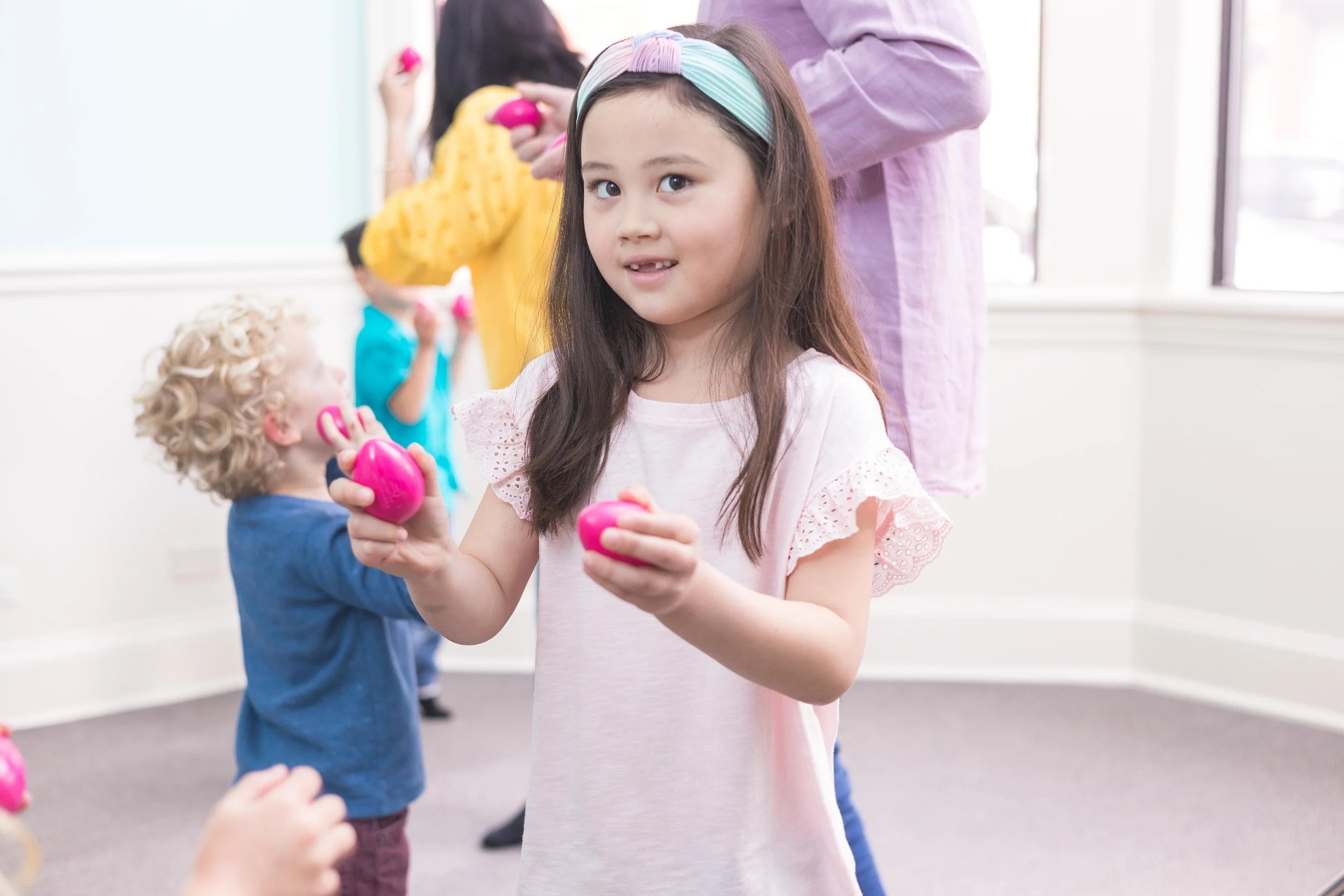 Young girl in a pink dress and pastel headband holding pink plastic eggs at an egg hunt. Other children in the background also holding eggs, indoors with bright windows.