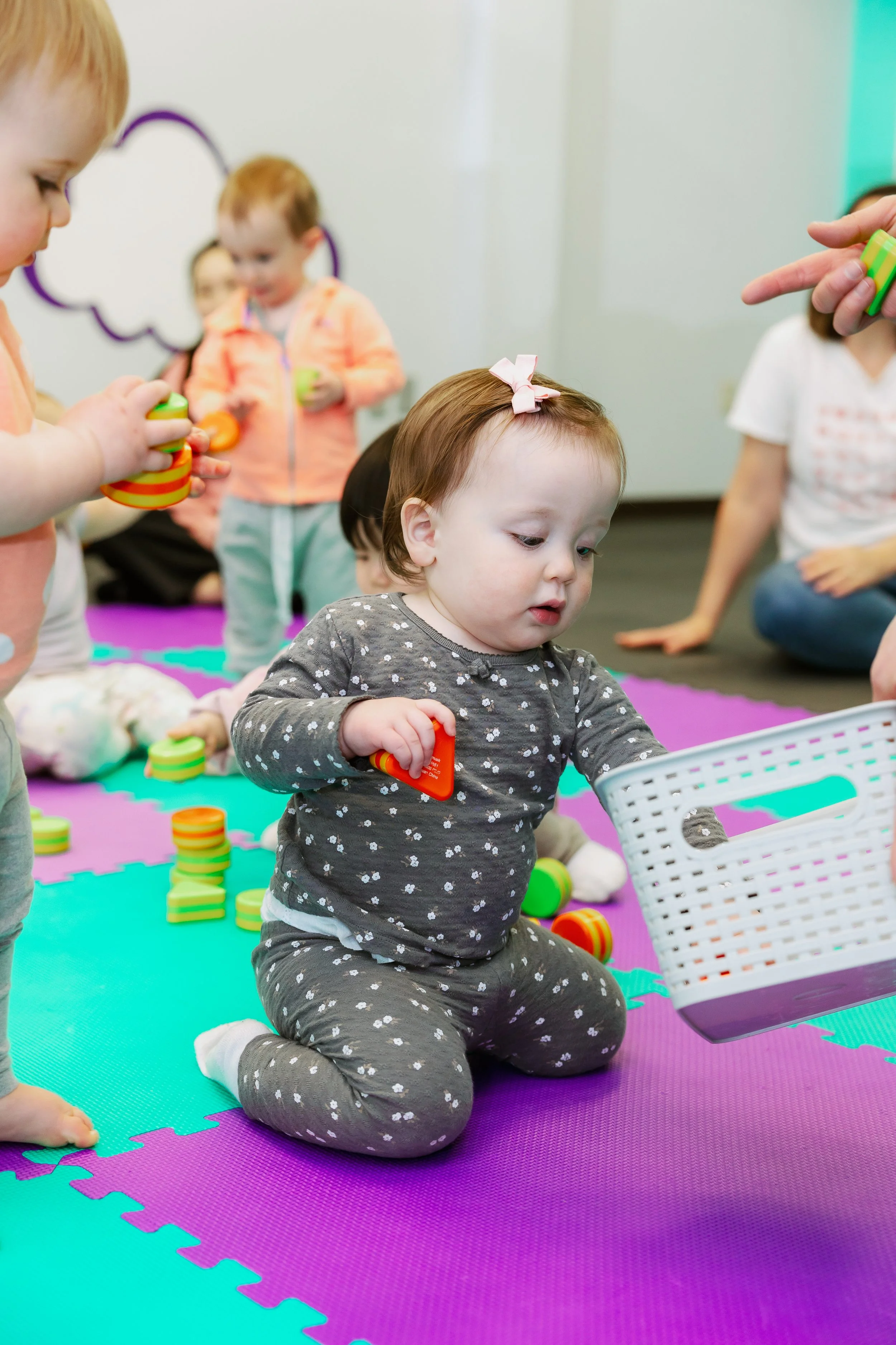 Young children participating in a play activity on a colorful foam mat, with one girl sitting and holding a toy, others in the background, some holding toys.