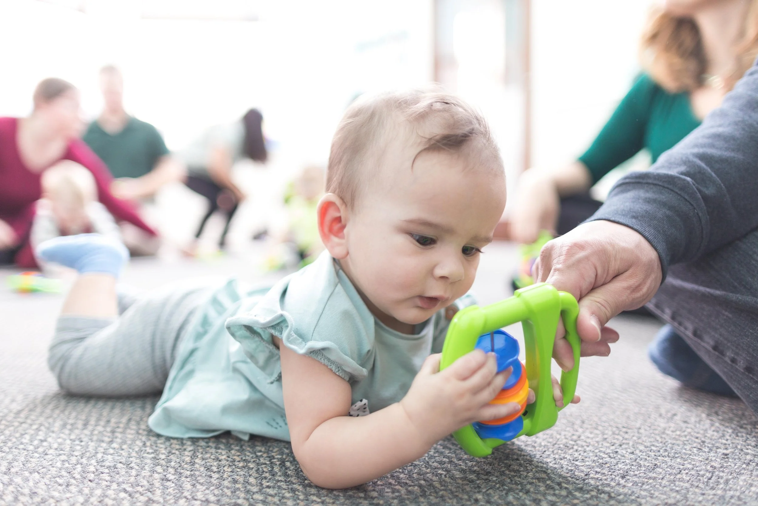 A young child lying on the ground playing with a colorful plastic toy, with adults in the background engaged in conversation.