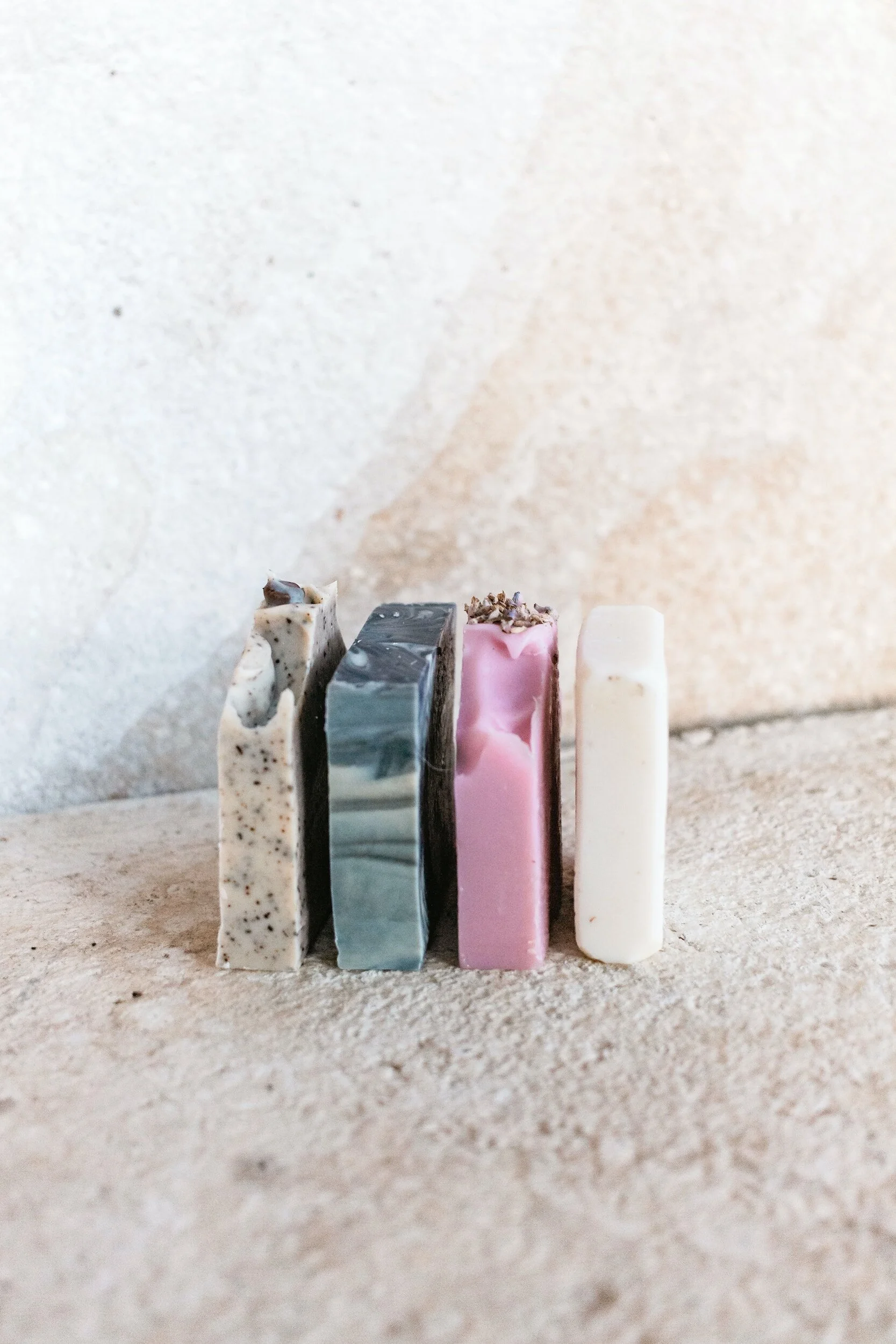 Four rectangular bars of soap with different colors and patterns, lined up on a textured surface against a plain wall.