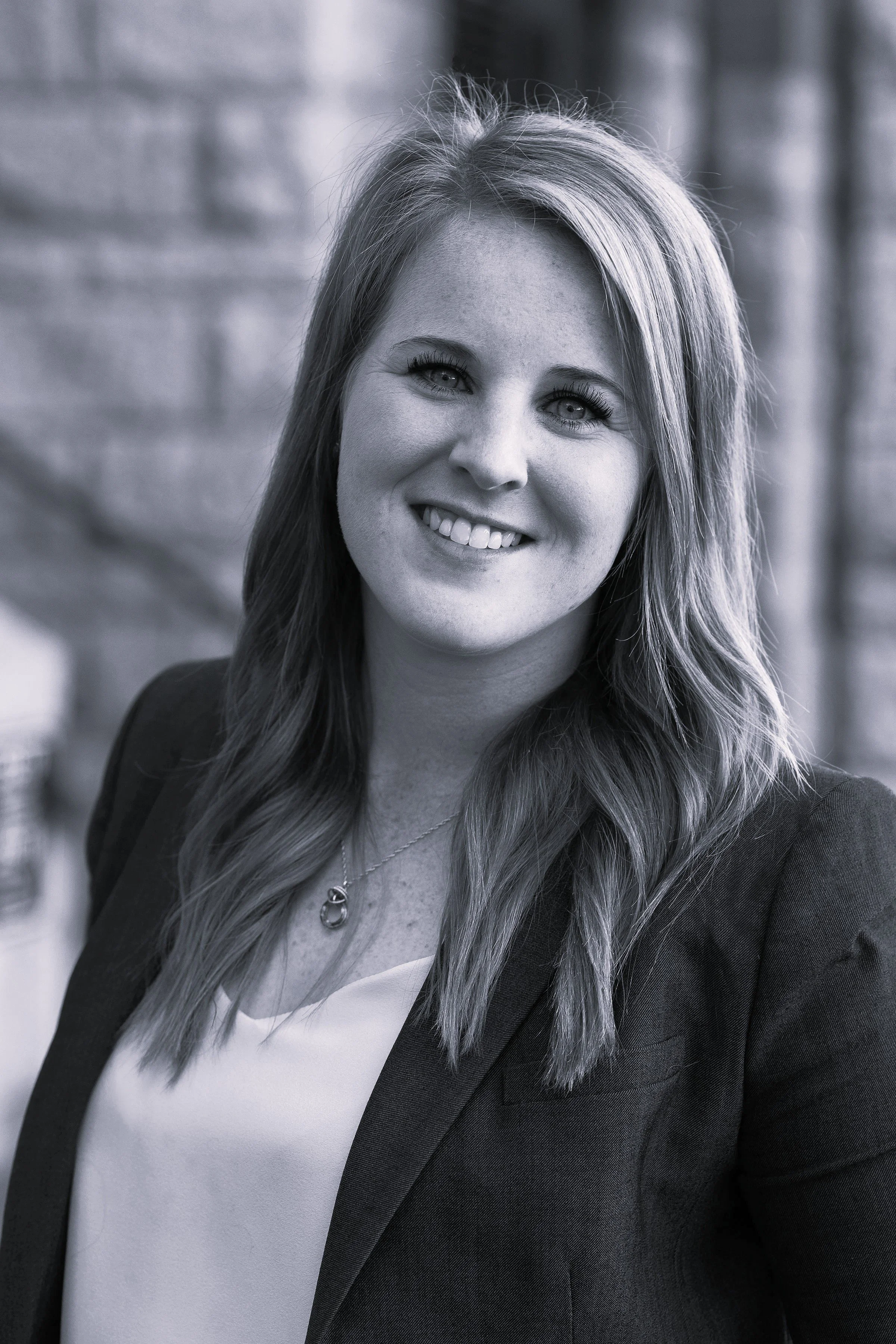 A smiling woman with long, wavy blonde hair wearing a navy blazer and white top, standing outdoors with a brick wall in the background.